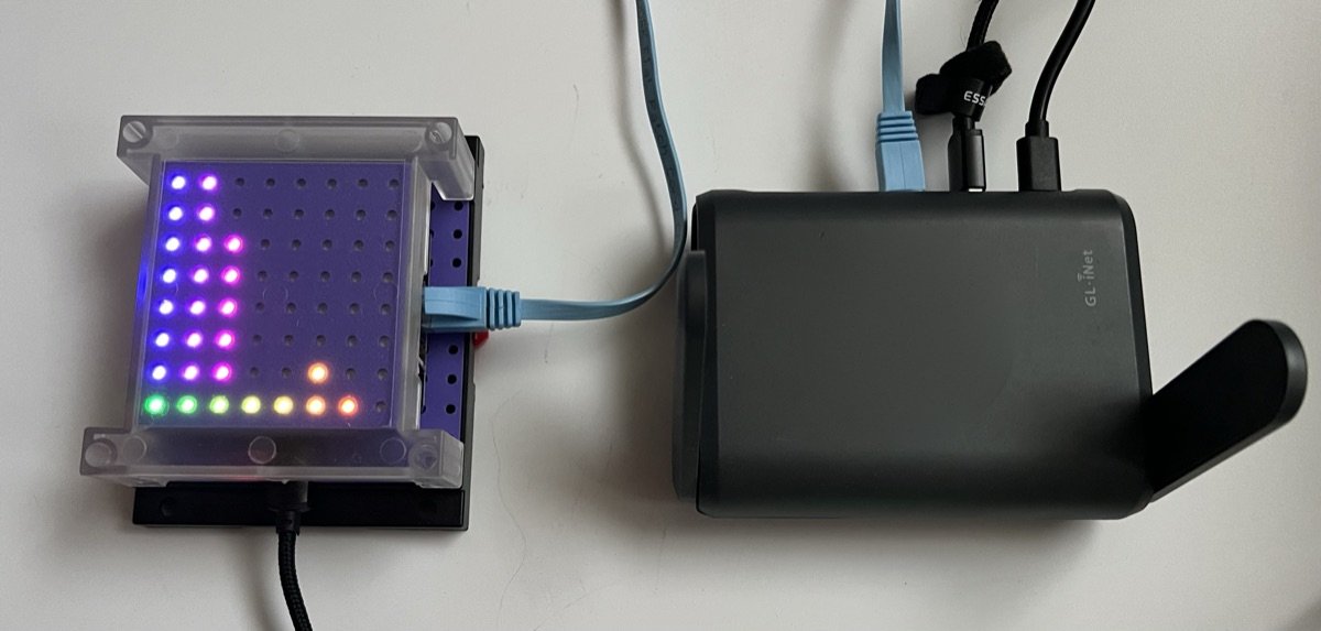 The ESP32-S31 CoreBoard in a clear case, its LED matrix showing what it hears, wired by Ethernet to a GL.iNet Slate AX travel router
